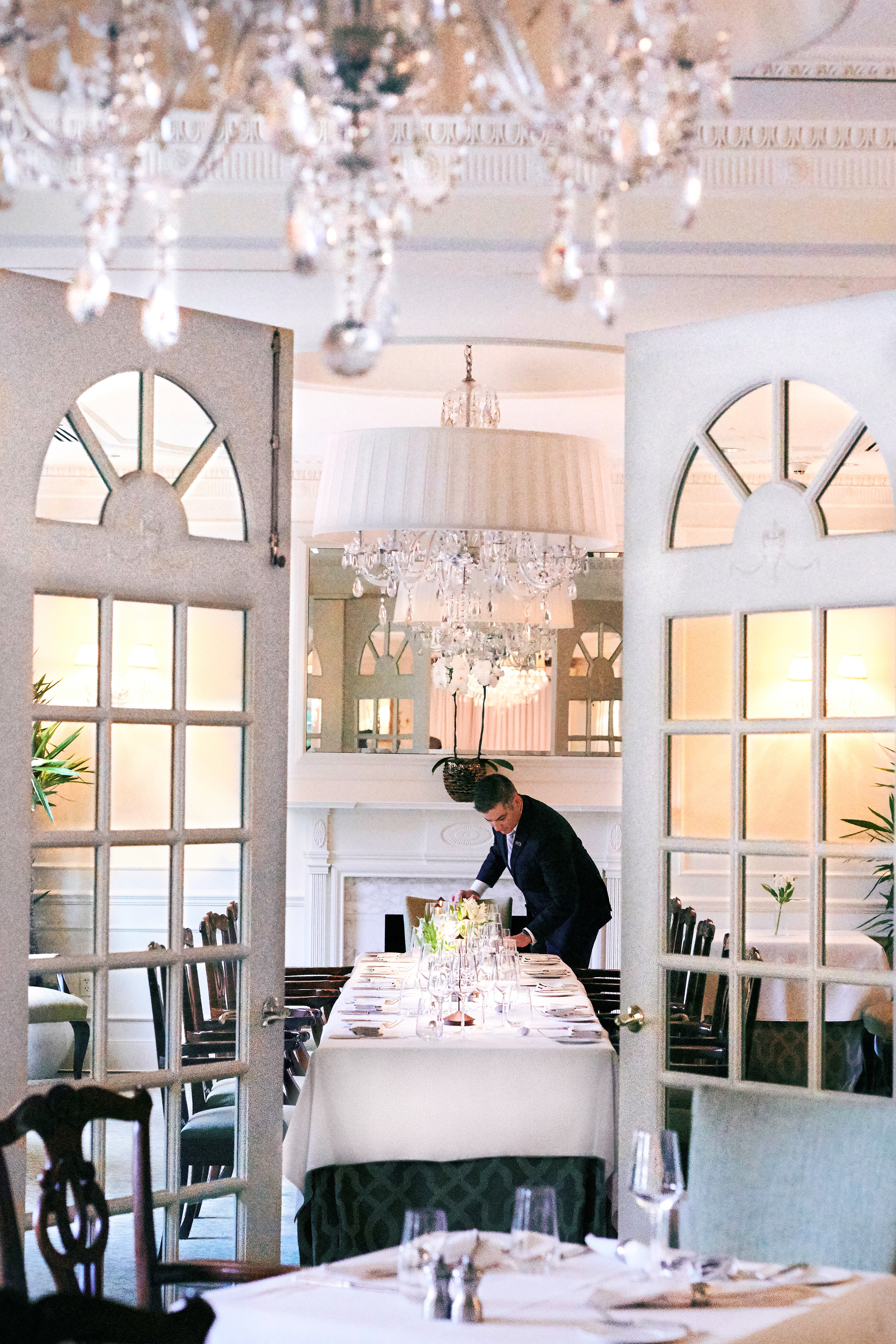 A man in a suit sets a table in a formal dining area with a chandelier and white tablecloth.