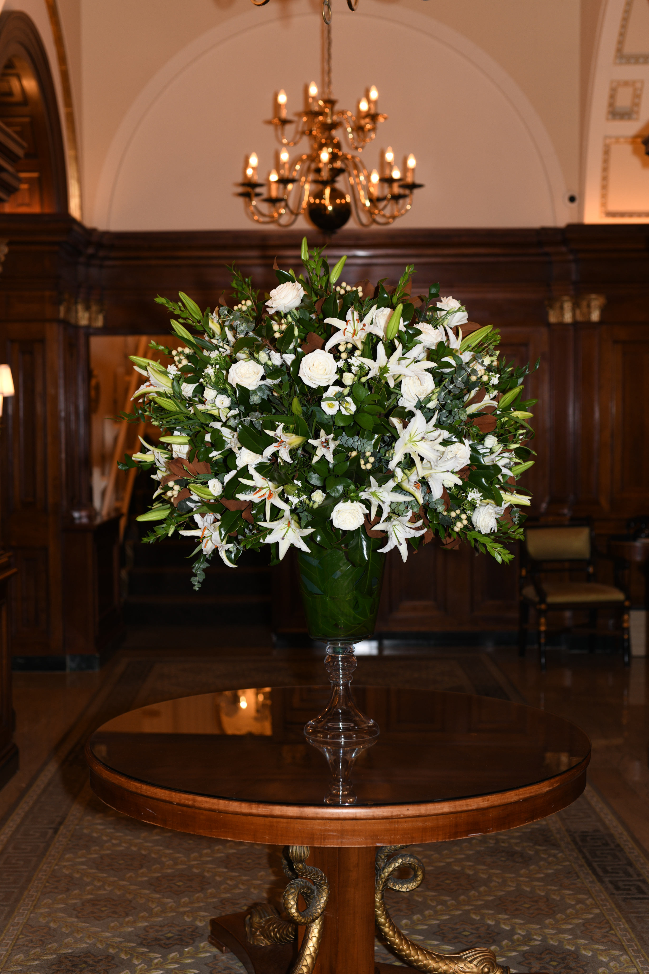 A large floral arrangement on a round wooden table in a lobby with a chandelier and wood paneling.
