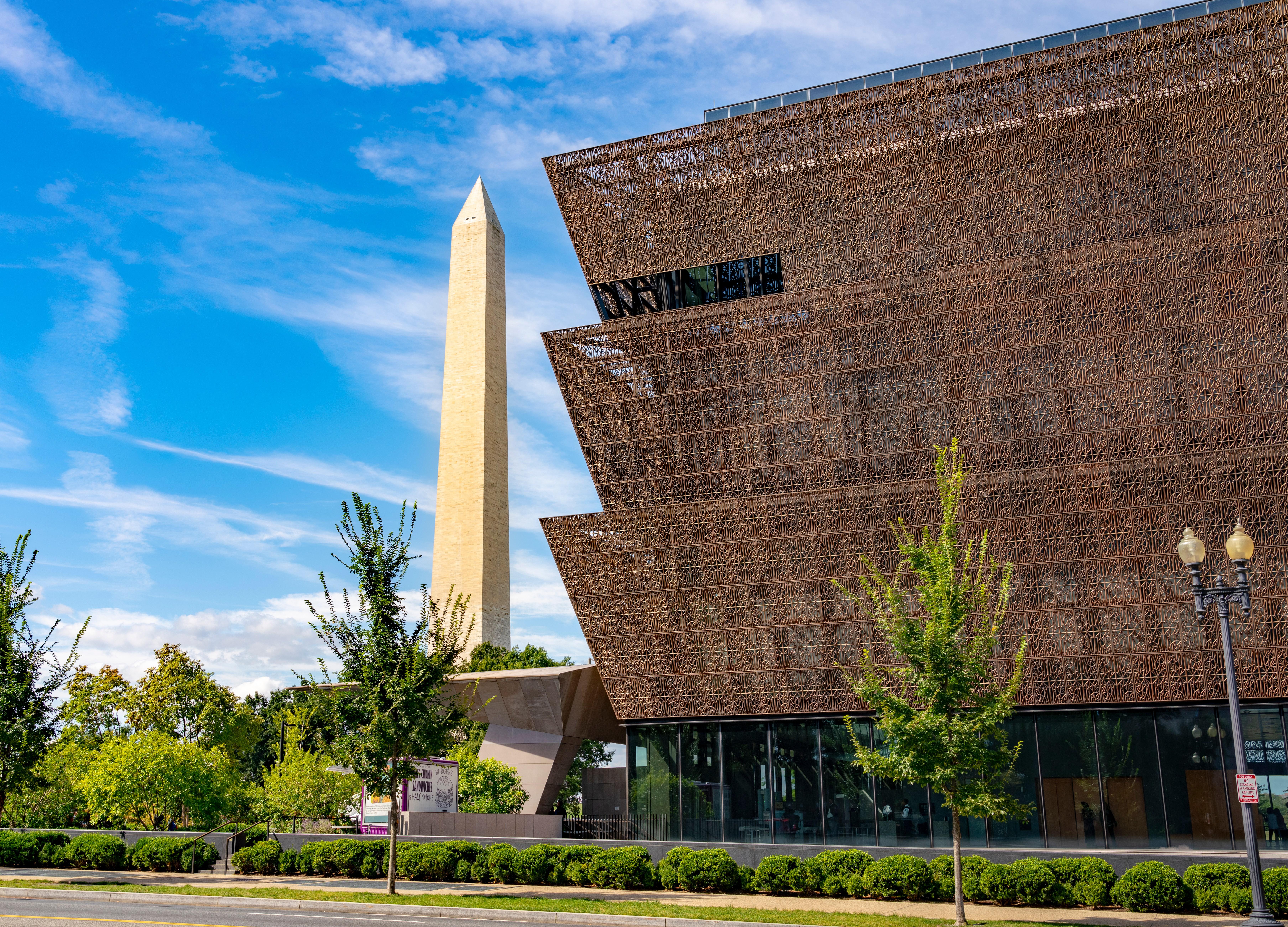 A large building with a brown facade and glass windows, surrounded by trees, with the Washington Monument in the background.
